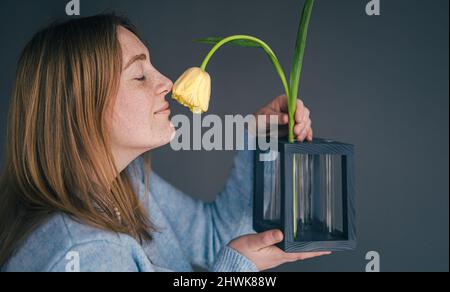 Niedliche junge Frau schnuppert eine Tulpe in einer Vase auf grauem Hintergrund. Stockfoto