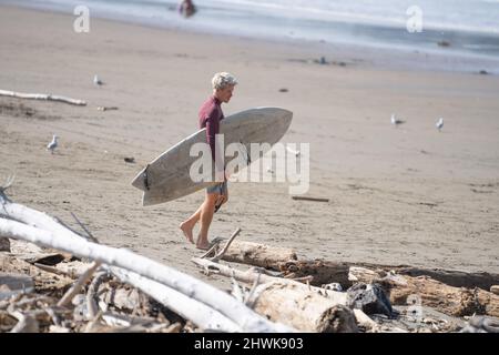 Der Surfer bringt das Brett zum Wasser, vorbei an Treibholz, das am Strand gewaschen wurde Stockfoto