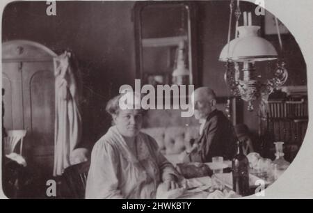 Ein antikes Foto über ein älteres Paar (Mann und Frau), das in ihrem wunderschönen Wohnzimmer in ihrem schönen Zuhause sitzt. Sie haben einen riesigen Spiegel auf der Rückseite und einen reich verzierten neobarocken Kronleuchter auf der rechten Seite des Bildes, einen großen Kleiderschrank (mit einem Bademantel) auf der linken Seite des Bildes. Auf dem Schreibtisch befindet sich eine große Glasflasche (Wein oder Mineralwasser), nur neben der Dame. Schönes Zuhause. Zeitraum: 1900er, Quelle: Originalfoto. . ADDITIONAL-RIGHTS-CLEARANCE-INFO-NOT-AVAILABLE / Kunstsammler und Heim Stockfoto