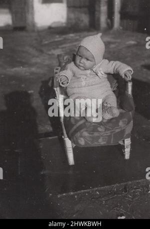 Antikes monochromes Foto über ein niedliches Kleinkind, das in einem Armstuhl sitzt und die Anf seiner/ihrer Mutter beobachtet, gab keinen Knacken über die Kamera. Sie/er hat ein schönes warmes Strickkleid mit Hut. Zeitraum: 1930s. Quelle. Originalfoto. ADDITIONAL-RIGHTS-CLEARANCE-INFO-NOT-AVAILABLE Stockfoto