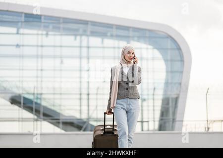 Flugreise und Geschäftskonzept. Hübsche Reisende Touristen-arabische Frau in Hijab, die Koffer und Telefon trug und in der Nähe des modernen Flughafenterminals ging Stockfoto