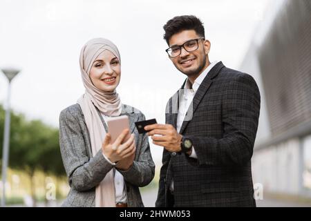 Zwei multiethnische Geschäftsleute, die online im Freien Finanztransaktionen durchführen. Muslimische Dame in Hijab Ihr arabischer Kollege hält per Smartphone für bargeldlose Zahlungen eine Kreditkarte. Stockfoto