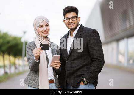 Zwei multiethnische Geschäftsleute, die online im Freien Finanztransaktionen durchführen. Muslimische Dame in Hijab Ihr arabischer Kollege hält per Smartphone für bargeldlose Zahlungen eine Kreditkarte. Stockfoto