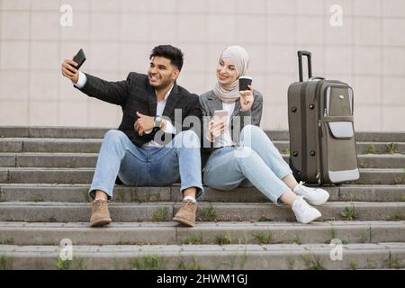 Angenehm lächelnder Inder und hübsche muslimische Dame im Hijab, die mit Reisekoffer auf der Treppe sitzt und Selfie macht. Ein Geschäftspaar sitzt auf der Treppe vor dem Flughafen. Stockfoto