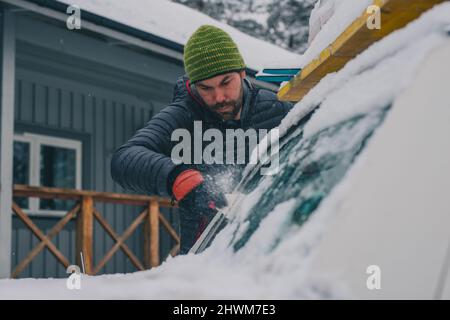 Ein junger modischer Mann putzt an einem kalten Morgen Autofenster mit einem Schaber. Person, die Handschuhe, Hut, technische Jacke und Fensterschaber zum Reinigen verwendet Stockfoto