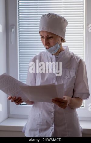 Die Ärztin steht in der Nähe des Krankenhausfensters und schaut durch die Dokumente. Stockfoto