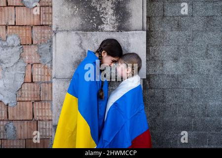 Kleine Mädchen mit ukrainischen und russischen Fahnen vor einer durch Bomben zerstörten Mauer. Ukraine russland Konflikt 2022 Eskalation. Konzept des Friedens Stockfoto