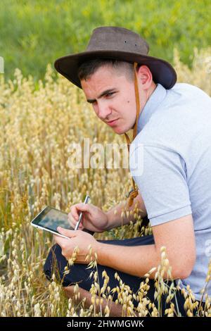 Digitale Farm. Porträt eines Bauern, der im goldenen Weizenfeld sitzt und auf dem Tablet scrollt. Junger Mann mit Cowboyhut auf dem Feld, der Weizenernte untersucht. Hafer Stockfoto