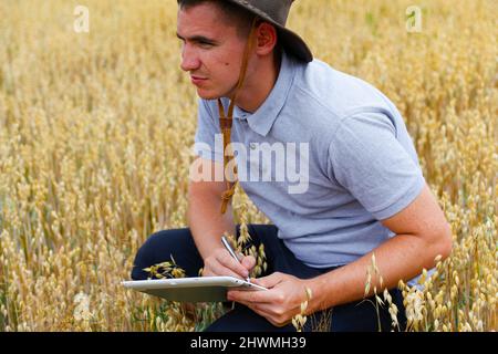 Digitale Farm. Porträt des Bauern sitzen in Gold Weizenfeld mit Tablet. Junger Mann mit Cowboyhut auf dem Feld, der Weizenernte untersucht. Hafer Getreideprodukte Stockfoto