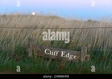 Warnschild am Cliff Edge am Beachy Head bei Eastbourne, Sussex, Großbritannien, wenn die Sonne untergeht. Stockfoto