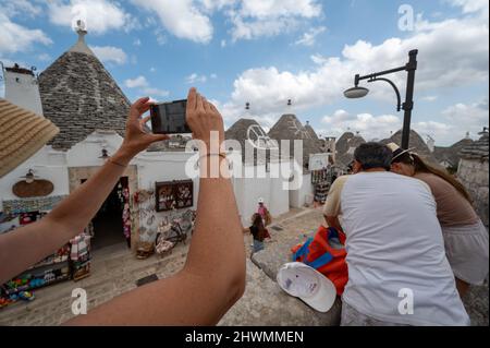 Alberobello, Apulien, Italien. August 2021. Herrliche Aussicht auf das eindrucksvolle historische Dorf. Eine Frau mittleren Alters fotografiert mit ihrem Mobiltelefon. Stockfoto