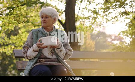 Kaukasische ältere obdachlose Frau, die auf einer Bank sitzt und eine Plastikschüssel mit heißer Suppe hält, die ihr von Freiwilligen gegeben wurde. Hochwertige Fotos Stockfoto