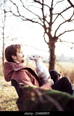Fröhlicher junger Mann mit Jacke sitzt draußen mit Hund auf dem Schoß in Sonnenschein Stockfoto