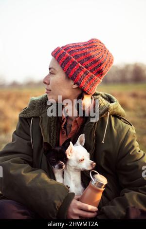 Gemischte ethnische Zugehörigkeit nicht-binäre Person sitzt im Feld in der Sonne mit zwei Hunden Stockfoto