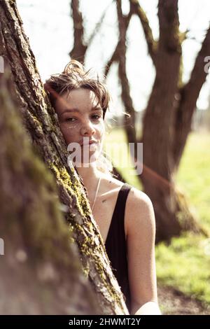 Junge Frau mit Sommersprossen lehnt sich an einen Baum mit Sonnenscherben in der Natur Stockfoto