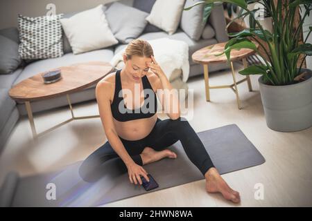 Schwangerschaft und Technoloy unterstützten ein gesundes Lebensstilkonzept. Fröhliche glückliche Schwangere mit Smartphone-Anwendung beim Training auf Yoga-Matte Stockfoto