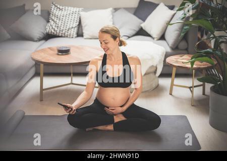 Schwangerschaft und Technoloy unterstützten ein gesundes Lebensstilkonzept. Fröhliche glückliche Schwangere mit Smartphone-Anwendung beim Training auf Yoga-Matte Stockfoto