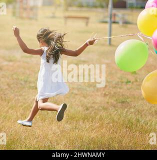 Glücklich hüpfend. Rückansicht eines kleinen Mädchens, das mit einer Reihe von Luftballons draußen durch ein Feld läuft. Stockfoto