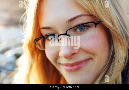 Geheimes Lächeln. Junge blonde Frau mit einer Brille, die die Kamera anlächelt. Stockfoto