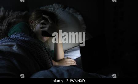 Ein abgedunkeltes Zimmer mit einem Mädchen im Bett auf ihrem Handy und Laptop. Konzept für Social Media, Mobbing im Internet, Schlaflosigkeit oder Bildschirmabhängigkeit. Stockfoto