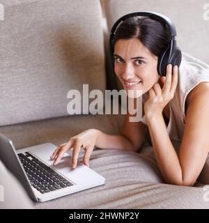 Im digitalen Zeitalter in Kontakt bleiben. Aufnahme einer jungen Frau auf einem Sofa, das Kopfhörer trägt und einen Laptop benutzt. Stockfoto