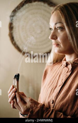 Frau meditiert mit palo santo im Studio und reinigt den Raum mit Rauch. Erwachsene Person, die zu Hause brennende spirituelle Räucherungen verwendet Stockfoto