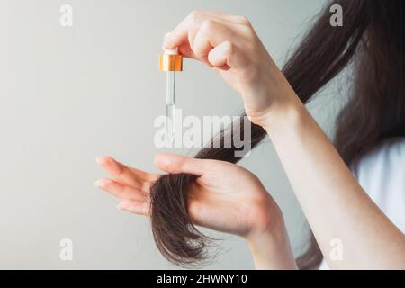 Eine Frau mit dunklem Haar trägt mit einer Pipette ein kosmetisches Produkt an die Haarenden auf. Nahaufnahme der Hände. Grauer Hintergrund. Das Konzept der Haare Stockfoto