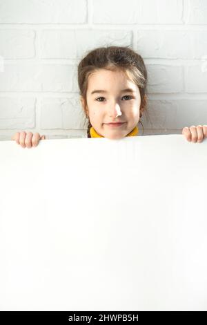 Ein trauriges Mädchen hält ein weißes Blatt Papier - Nachbau für Werbung, Slogan, Inschrift. Kopierraum liegt in den Händen der Kinder, das Kind ist verärgert und versteckt Stockfoto
