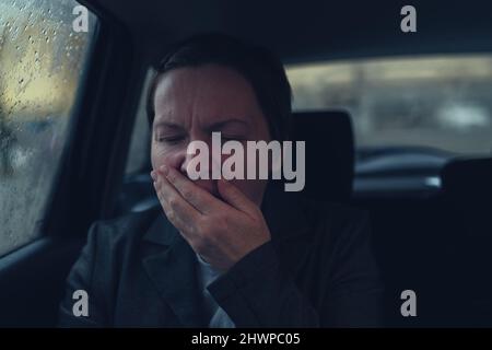 Müde erschöpfte Geschäftsfrau, die auf dem Rücksitz des Taxifahrzeugs auf dem Heimweg von der Arbeit gähnt, selektiver Fokus Stockfoto