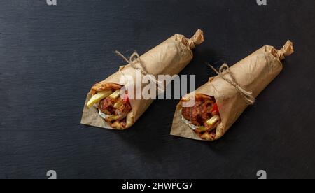 Gyro Pita Shawarma auf schwarzer Steinplatte. Griechische Lebensmittel, Fleisch in Scheiben, Kartoffeln, Tomaten und Zatziki, Papierverpackung, Draufsicht. Stockfoto