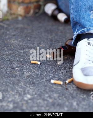 Die Jugend ist schlecht geworden. Nahaufnahme einer Person, die neben weggeworfenen Zigarettenkippen und Bierflaschen sitzt. Stockfoto