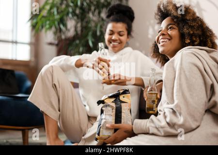 Zwei glückliche junge afrikanische Freundinnen sitzen auf einer Couch, trinken Bier und essen Chips zu Hause Stockfoto