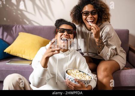 Zwei glückliche, multiethnische Freundinnen sitzen zu Hause auf einer Couch und halten sich lachend Snacks vor dem Fernseher Stockfoto