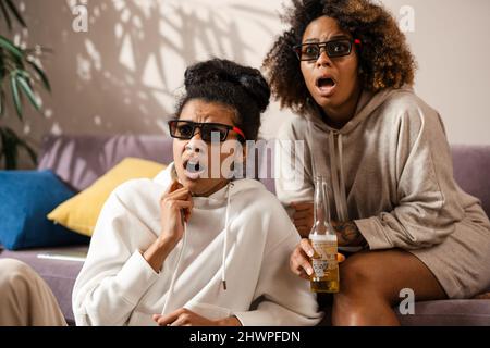 Zwei schockierte multiethnische Freundinnen, die zu Hause auf einer Couch sitzen und Snacks in 3D Gläsern beim Fernsehen sehen Stockfoto