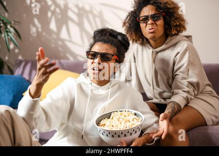 Zwei schockierte multiethnische Freundinnen, die zu Hause auf einer Couch sitzen und Snacks in 3D Gläsern beim Fernsehen sehen Stockfoto
