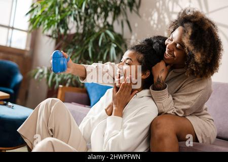 Zwei glückliche junge afrikanische Freundinnen machen Selfies mit einer Sofortkamera, die zu Hause auf einer Couch sitzt Stockfoto