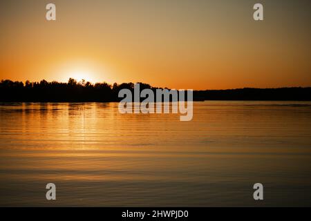 Ruhiger, gewellter See mit Sonne, die fast unter dem Horizont untergeht und die Wasseroberfläche mit seinen Strahlen und Bäumen am Ufer im Hintergrund bedeckt. Erstaunliche Aussicht voll Stockfoto