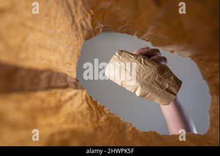 Eine Hand legt ein in Papier verpacktes Mittagessen in eine Papiertüte. Die Hand eines reifen Mannes hält das Essen mit ihm über den offenen braunen Beutel. Von unten nach oben geschossen. Clo Stockfoto