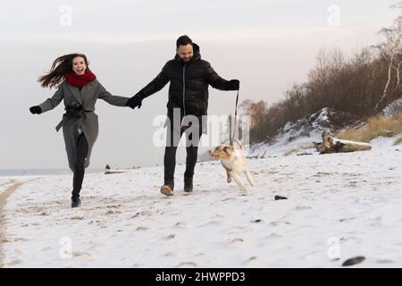 Glückliches junges Paar, das bei Sonnenuntergang mit Hund auf Schnee läuft Stockfoto