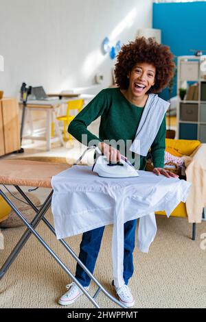 Fröhliche junge Afro-Frau bügelt Hemd an Bord im Wohnzimmer Stockfoto