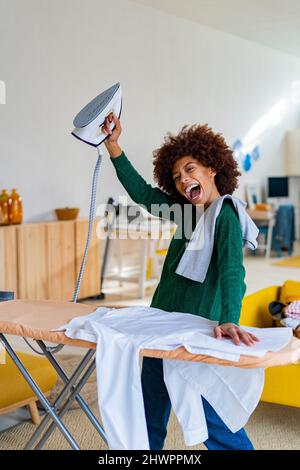 Fröhliche junge Afro-Frau, die Eisen an Bord mit Hemd im Wohnzimmer hält Stockfoto