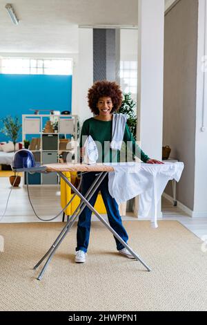 Lächelnde junge Afro-Frau mit Hemd an Bord, das im Wohnzimmer Bügeleisen hält Stockfoto