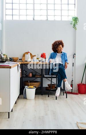 Nachdenkliche junge Afro-Frau, die auf dem Hocker sitzt und sich zu Hause auf dem Küchentischtisch lehnt Stockfoto