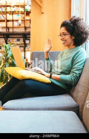 Lächelnde Geschäftsfrau winkt bei einem Videoanruf durch den Laptop auf dem Sofa Stockfoto