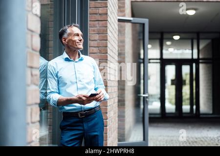 Selbstbewusster, reifer Geschäftsmann, der sich vor dem Bürogebäude am Fenster stützt Stockfoto