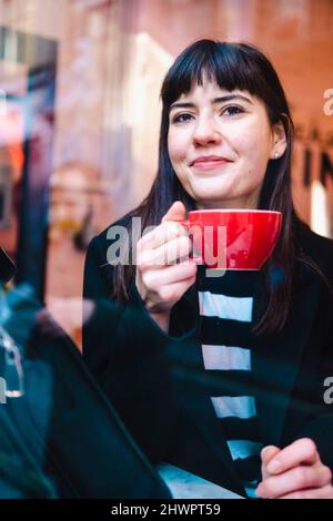 Lächelnde junge Frau mit Kaffeebecher und Blick durch das Fenster Stockfoto