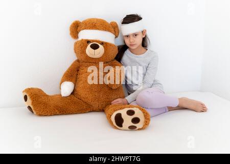 Verband auf Mädchen mit Teddybär sitzt vor der weißen Wand Stockfoto