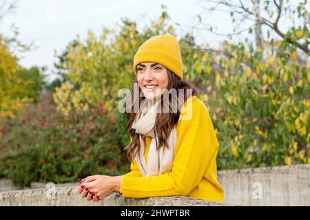 Junge Frau mit gelbem Pullover und Strickmütze, die im Park an der Wand gelehnt ist Stockfoto