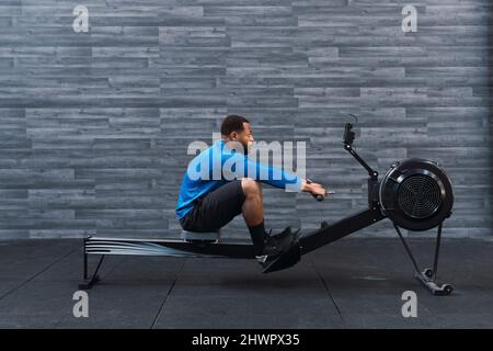 Konzentrierter Sportler trainiert auf dem Rudergerät vor der Wand im Fitnessstudio Stockfoto