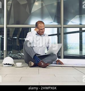 Er ist immer im Arbeitsmodus. Aufnahme eines fokussierten professionellen Architekten, der draußen auf dem Boden sitzt, während er an einem Laptop arbeitet. Stockfoto